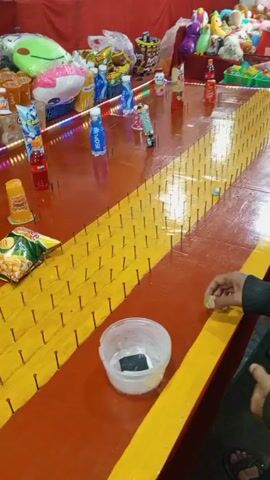 The video shows a game at a fair or carnival. A red and yellow table is covered with rows of nails. A person's hand pushes a token across the table, aiming for a plastic container of water with a phone inside. Various food and drink prizes are on the table, including chips, juice, and soda. Other plush toys and snacks are on display in the background.