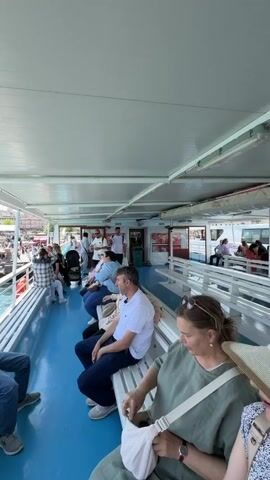 The video shows the interior of a covered passenger ferry with people seated on benches. The floor is blue, and the ceiling is white. On the left side of the ferry, there is a railing and a view of other boats and a cityscape. A young person in a white shirt and blue jeans is sitting on the bench, looking at their phone. Several other passengers are visible, some facing forward and others looking out at the water. The water is a clear turquoise color. On the right side, the view opens up to show a mosque with minarets and more boats. Further along, a bridge with traffic is visible. The overall atmosphere appears to be one of casual travel during the daytime.