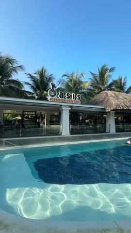 A still shot shows a resort-style swimming pool in the foreground with a fitness center in the background. The fitness center has a thatched roof and a sign that reads "Oasis Fitness Center." Several people are visible swimming in the pool. Palm trees surround the area, and the sky is clear and blue.