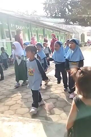 A group of young children, mostly girls wearing hijabs, are gathered outdoors in front of a green building. Some are wearing matching blue hijabs and dark athletic wear. One girl in the foreground wears a light blue tunic with gray and black stripes and dark pants. Another girl in a white hijab and green dress stands to the left. The children appear to be engaged in some sort of organized activity or exercise, with some raising their hands or moving their bodies. The ground is paved with hexagonal stones. A person in a red shirt is visible further back near the building, possibly an adult instructor or supervisor. The sun is shining, casting shadows.