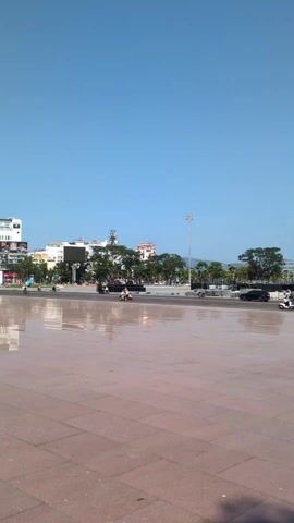 The video presents a wide shot of a city square or plaza on a clear, sunny day. The foreground is dominated by reddish-brown square pavers, reflecting the sky, creating a mirroring effect on the surface. In the middle ground, a street with a few moving motorcycles and a dark-colored vehicle can be seen. Beyond the street are buildings of varying heights and architectural styles. There are trees interspersed amongst the buildings, some appearing to be palm trees. The sky is a bright, cloudless blue. A tall lamppost stands out against the urban landscape.