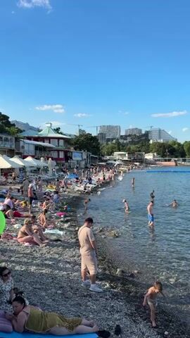 The video shows a crowded beach on a sunny day. Many people are sunbathing on the pebble beach, some are sitting on chairs, and others are playing in the shallow water. There are several buildings and structures along the coastline, including what appear to be cafes or shops. In the background, there are more buildings, possibly hotels or residential complexes, and construction cranes are visible. The sea is calm and clear. A few children are playing in the water near the shore. The overall atmosphere is one of a bustling summer vacation spot.