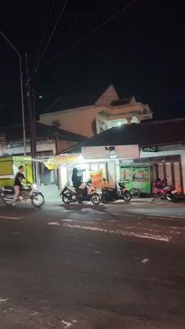 The video shows a street scene at night. The street is paved and has white markings. On the right side of the street, there are several parked motorcycles. Further down the street, there are buildings with awnings and lights, suggesting shops or food stalls. Some people are visible standing and walking around. In the center of the frame, a person is riding a motorcycle, which is blurred due to motion. A streetlight is visible on the left side of the frame. The overall atmosphere is that of a typical street scene in a commercial area at night.