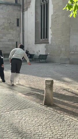 Two men are seen walking away from the camera in a sunny outdoor space, which appears to be a courtyard. One man is wearing a light-colored sweater and dark pants with a white stripe down the side, while the other is wearing a black tracksuit. They are walking on a cobblestone surface towards a gravel area where several metal balls are scattered. In the background, a large stone building is visible, featuring a tall, arched window. Two park benches are positioned against the building's wall, and a person is sitting on one of them. A stone post stands near the foreground. The overall scene suggests a relaxed, casual atmosphere.