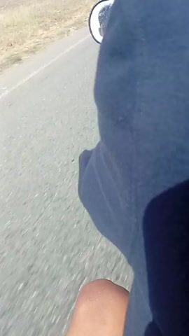The video depicts a person riding a motorcycle on a paved road under a clear, sunny sky. The rider, seen from behind, is wearing a dark blue long-sleeved shirt. The view includes the motorcycle's dashboard, handlebars, and side mirror. The side mirror reflects the rider's upper body and the surrounding environment. The road has some white lines, and the surrounding area includes fields, buildings, and power lines. Several vehicles are visible in the distance, including a truck with a crane. The motorcycle is moving at a moderate speed.