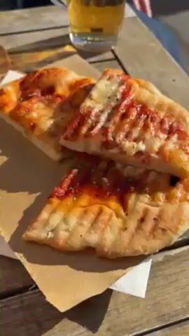 The video shows a wood table with an open-faced pizza and a glass of beer. The camera pans back to show a couple sitting at a table overlooking the sea on a sunny day. The ocean is sparkling, and there are trees along the sidewalk.