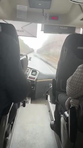 The video shows the interior of a Higer bus, viewed from behind the first row of seats. Rain is visible on the windshield, which also has a large crack. The dashboard and steering wheel are visible, along with a digital clock displaying "8:26". The bus is traveling on a road lined with trees. Two individuals are partially visible in the first row seats, one wearing a dark jacket and the other wearing a quilted cream-colored garment. The view is slightly shaky, suggesting the bus is in motion.