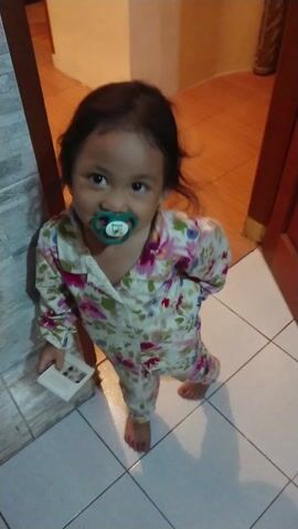 The video captures a baby standing indoors, looking directly up at the camera. The child has dark hair and wide, expressive eyes. They are wearing a floral-patterned long-sleeved outfit with various shades of pink, purple, green, and white. A green pacifier is in the baby's mouth, featuring the letter "B" prominently on its front. In their right hand, the baby is holding a small white rectangular object, possibly a phone or a remote control. The flooring in the foreground is light-colored tile, transitioning to a wooden or wood-patterned floor in the background. The scene depicts a casual moment in a home setting.