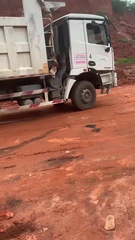 A white dump truck with a large trailer is parked on a reddish-brown dirt road. The truck has the number 4 on the driver's side door and a pink sticker with white text that reads "MAYBE SOULTIONS". A person wearing a patterned blue and white outfit is walking away from the truck in the background. The road appears to be a construction site or a mine, with piles of red dirt visible in the background. The ground is uneven and dusty, with tire tracks and footprints visible. The weather appears overcast.