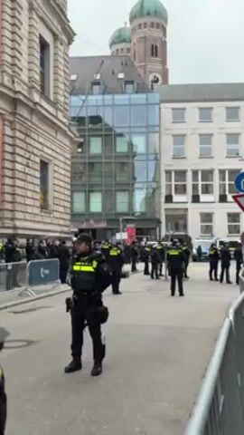 The video shows a street in what appears to be a European city.  Numerous police officers in black uniforms with fluorescent yellow accents are standing along the street, facing forward.  Barriers are set up on both sides of the street. Behind the barriers on the left, a small crowd of people in suits is visible. Buildings of various architectural styles line the street, including one modern building with a reflective glass facade. In the distance, a building with a green dome is visible.  A loud, unidentifiable noise is heard.