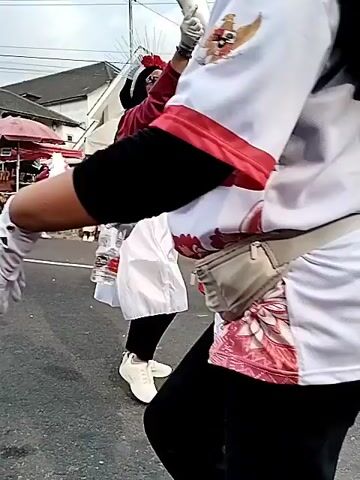 The video shows a parade or festival on a paved road. Several people are visible, some in costumes. In the foreground on the left, a person in a red and white outfit with white gloves is walking away from the camera. To their right, a person in a black and white marching band-style uniform with a pink tutu is walking towards the camera. Further in the background, people dressed in traditional Indonesian attire are visible, along with spectators. There are tents and stalls set up along the side of the road, and flags, including Indonesian flags, are flying. The environment appears to be a street or public area.