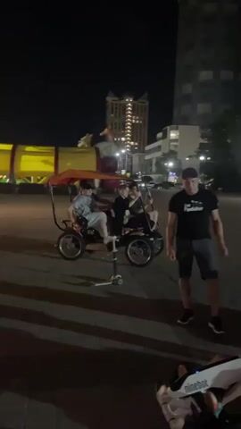 At night, a group of people are gathered around several small motorized vehicles, possibly go-karts or similar recreational devices. Two of the vehicles are in the foreground, white and black with the "ninebot" logo visible. Several people are interacting with these vehicles. In the background, a four-wheeled, pedal-powered cart with a red canopy is moving away from the camera, carrying at least three passengers. The scene takes place on a paved area, likely a plaza or promenade, with buildings and streetlights visible in the background. The atmosphere appears casual and recreational.