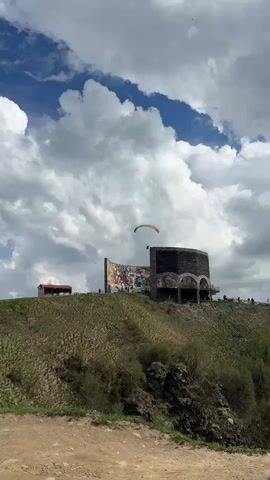A paraglider with a yellow and red canopy maneuvers in the sky above a grassy hill. On the hill sits a large, curved structure adorned with a colorful mosaic mural depicting numerous figures in various poses. To the left of the mosaic structure, a smaller red building with a dark roof is visible. Several people are standing on the edge of the hill near the mosaic. The sky above is filled with dramatic, white, and gray clouds. The foreground shows a dirt path. The paraglider is in motion, suggesting an active flight.
