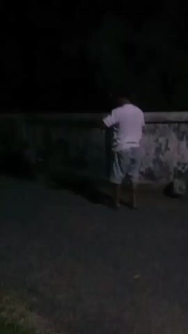 The video begins with a blurry, dark shot of a person in a light-colored t-shirt and patterned shorts standing near a wall. The camera then pans to the right, showing a dark scene with some faint lights in the distance. The video then shows a person carrying a baby, walking along a street at night. Several other people are visible in the background, some sitting or lying on the ground. There are streetlights casting a dim glow on the scene. The video ends with a person holding a white flag. The overall scene appears to be outdoors, possibly a street or gathering, at night.