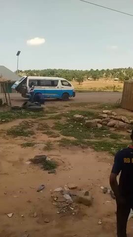 A full outdoor shot shows a white and blue van parked on the left side of a dirt road, with its back door open. A person in a blue shirt is seen near the back of the van, possibly working on it. Several tires are scattered on the ground near the van. In the foreground, a person wearing a dark blue shirt with "RB LE" visible on the back stands facing away from the camera. The ground is dusty and covered with sparse green patches and rocks. In the background, there's a light-colored building or structure on the left and a dirt road extending into the distance, with green foliage and distant structures under a clear sky. Another person in orange walks from the right side of the screen towards the center.