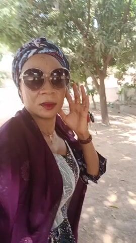 A woman stands outdoors, taking a selfie in front of a tree. She is wearing a colorful head wrap, large round sunglasses, a dress with a beaded bodice and a maroon shawl. She is smiling and holds her hand up to her face in a waving gesture. The background includes trees, a car, and a grassy area. The scene appears to be taken during the day.