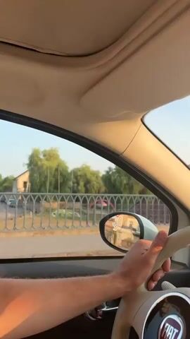 This media file captures a first-person perspective from inside a Fiat car, driving through a town. The driver's hand is visible on the steering wheel. Through the windshield, a street with buildings and trees can be seen. In the distance, a church spire is prominent, and there are also fountains visible on the street. The time of day appears to be late afternoon or early evening, indicated by the warm, golden light bathing the scene. The sky is clear and blue, suggesting pleasant weather.

The car's interior is also visible, including the dashboard, rearview mirror, and a sun visor. The Fiat logo is clearly visible on the steering wheel. The surrounding environment suggests a European town setting, with architecture that appears to be historic. The presence of a bridge and a river or canal can be inferred from the railing and water features. The overall mood is one of a leisurely drive through a picturesque town.

There is no visible text in the media file other than the "FIAT" logo on the steering wheel. The location context provided is Baccarat, France.