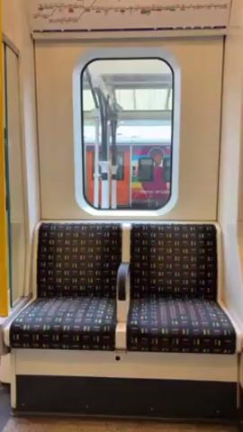 The video shows an empty two-seat bench inside a train car. The seats have a colorful geometric pattern. A small, rectangular window above the seats offers a view of a train with colorful graphics on its side, including what appears to be the phrase "Mayor of London". The walls of the train car are a light, neutral color. A map of some kind is visible along the top edge of the frame. The lighting is bright and artificial.