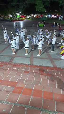 The scene, possibly at night, shows a marching band consisting of roughly 20 people assembled on a square paved with large rectangular tiles. The band members are dressed in white uniforms and hats, and they're holding instruments like trumpets and drums. The drums bear the colors of the Venezuelan flag. A young girl, wearing a red shirt and jeans, walks into the frame, down a flight of red brick stairs towards the band. There are other people scattered in the background, some sitting on benches. A green food cart stands near some white "YO" block letters. The environment appears to be a public square.
