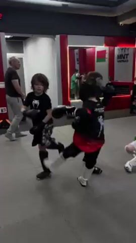 The video shows a group of young children, likely between the ages of 5 and 8, practicing kickboxing in a gym. They are wearing boxing gloves and protective gear like shin guards and headgear. An adult male stands off to the side, seemingly observing or instructing them. The gym has a padded floor, mirrored walls, and red and white trim.  One child is seen sparring lightly with another. The overall atmosphere suggests a training session for young martial arts enthusiasts.