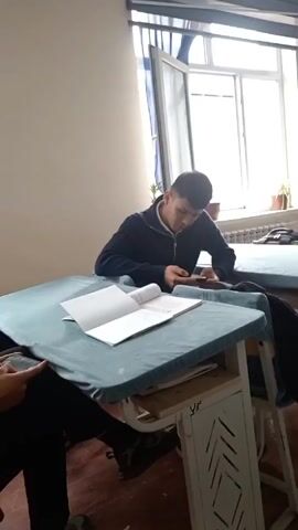Two young men sit at desks in a classroom, both looking at their phones and tapping the screens. A partially open book lies on the desk between them. Sunlight comes in through a window in the background. One of the men coughs.