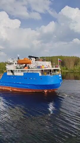 Here's a description of the video:

The video shows a blue and white river barge, named "Irina", moving across a body of water, likely a river or canal. The vessel leaves a wake behind it. The sky is blue with fluffy white clouds. The barge has a Russian flag displayed at its stern. The surrounding environment includes trees lining the banks of the waterway and power lines are visible in the background. The water reflects the sky.