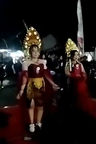 Two individuals, dressed in elaborate traditional attire with golden headpieces and flowing red fabrics, walk across a red carpet at night. They appear to be part of a procession or performance, with a crowd of onlookers visible in the background. The lighting is dim, suggesting an outdoor evening event.