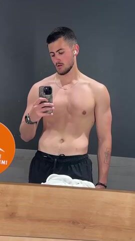 A young, light-skinned man with dark hair and a short beard takes a selfie in a mirror. He is shirtless, wearing black athletic shorts, a gold necklace, a watch, and wireless earbuds. He has a tattoo on his left forearm. He holds a smartphone with a camouflage case in his right hand, looking down at the screen. The background is a gray wall with a wood panel below it. An orange sign with white text is partially visible on the left side of the frame. A white cloth rests on the wood panel in front of the mirror.