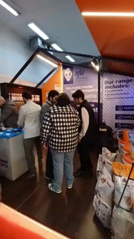 The video shows the interior of a commercial space, likely a trade show or exhibition booth. Several people are visible, including two men who are the focus of the shot. One man is wearing a black and white checkered shirt and jeans, facing away from the camera. The other man is wearing a black vest over a white shirt and dark pants. They appear to be engaged in conversation, standing near a table displaying promotional materials. The table features a stack of brochures or product catalogs with the words "PRODUCTS SELECTOR" visible on the cover. Behind them, a backdrop with the company logo "ayla" and the words "POLYURETHANE WATERPROOFING" is visible. The booth also has shelves displaying various products, including buckets and bags with labels. The lighting is bright, with fluorescent lights illuminating the scene. The overall atmosphere suggests a business setting focused on product demonstration and customer interaction.
