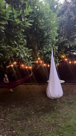 This media file depicts a backyard scene in Alakhadzi, Abkhazia, during what appears to be dusk. The setting is a grassy yard with lush green trees providing shade. String lights are visible, strung between trees and along a fence, casting a warm, ambient glow that suggests an evening atmosphere. The weather seems calm, with no visible rain or strong winds.

The primary objects in the scene are two hanging seats. On the left, a red hammock is suspended between two points, one of which is a tree trunk. On the right, a teardrop-shaped hanging seat, adorned with a grey and white chevron pattern, is suspended from a wooden frame. Behind the hanging seats, a fence runs horizontally across the frame. A person is visible in the hammock on the left, partially obscured by the fabric, appearing to be relaxing. The overall mood conveyed is one of tranquility and leisure, typical of a relaxing evening in a garden. The specific location cues, like the trees and the general layout of a backyard, are consistent with a residential setting. No visible text is present in the media file.