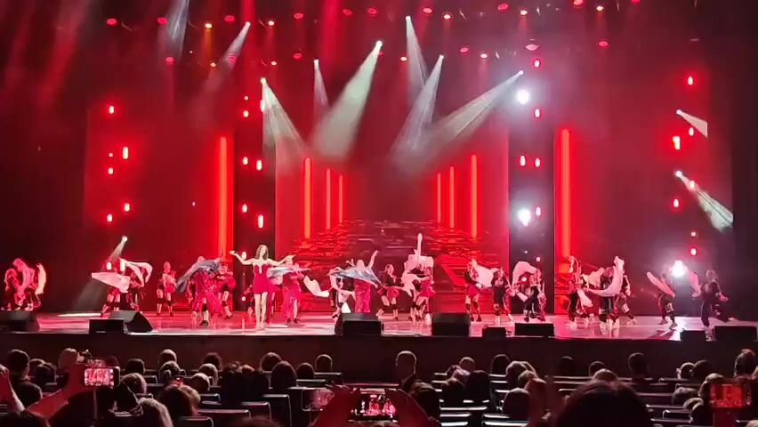 A group of dancers perform on a stage under red lights, with a group of people in the audience watching.