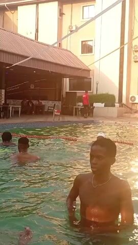 The video shows a group of young men swimming in an outdoor pool. Some are submerged up to their shoulders, while others are partially out of the water. One of the men is gesturing with his hand towards the camera. The pool area appears to be adjacent to a building with outdoor seating under a covered awning. The lighting suggests it is daytime.