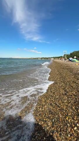 The video presents a serene, wide shot of a pebbly beach in Taman, Russia, on a bright, sunny day. Gentle, foamy waves lap rhythmically against the shore, which is covered in smooth, multi-colored pebbles. The sky is a vibrant blue with scattered white clouds. In the distance, along the coastline, what appears to be a small town or settlement can be seen. On the right side of the frame, further down the beach, a white lifeguard stand is visible. Several people are seen relaxing on the beach, some lying under colorful umbrellas, enjoying the tranquil atmosphere. The water appears clear, and the overall scene conveys a peaceful day at the beach.