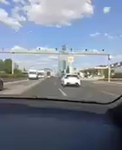 The video shows a view from inside a vehicle moving on a multi-lane highway. A white car is visible in front of the vehicle, and several other vehicles, including vans and cars, are on the road ahead and in adjacent lanes. To the right of the road, a Shell gas station sign with its iconic yellow and red logo is prominently displayed. Beyond the gas station, a modern, tall, glass-covered skyscraper dominates the skyline, with a distinctive stepped architectural design. Further in the distance, buildings and infrastructure suggest an urban or suburban environment. The sky is blue with scattered white clouds, indicating daytime.