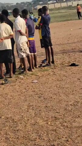 The video depicts a group of young men gathered on the edge of a dirt field. Some are standing and watching a soccer match that is taking place on the field. Others are sitting on the ground, also watching the game. The men are wearing casual clothing, including t-shirts, shorts, and sandals. Some of them have towels or bags with them. In the background, there is a small town or village with a hill or mountain visible in the distance. The sky is overcast.