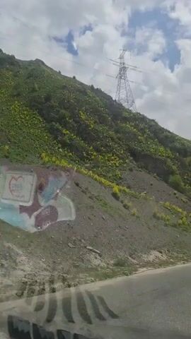 The video shows a view from inside a car driving on a paved road that curves slightly.  The road appears to be in a mountainous region. On the side of the road, there is a steep hill covered in green vegetation and small yellow flowers. Near the bottom of the hill, there is graffiti on a rock face. A tall electrical transmission tower stands on the hill. In the background, there are snow-capped mountains.  The sky is cloudy with patches of blue. A white car is seen briefly driving in the opposite direction.  The video includes some indistinct voices.
