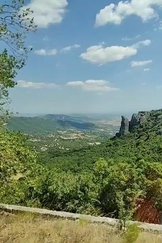 The video pans across a scenic landscape, revealing a vast valley dotted with trees and distant settlements under a bright blue sky with fluffy white clouds. In the foreground, lush green foliage frames the view, with a prominent, tall rock formation visible on the right side of the valley. The camera moves, showing more of the dense vegetation and the expansive vista, suggesting a natural environment, possibly in a mountainous or hilly region.
