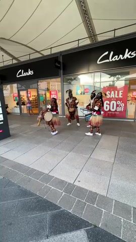 Four people in traditional African-style clothing and masks are performing on a paved walkway in front of a Clarks shoe store. They are playing drums and moving rhythmically. There is a sale sign visible in the store window offering up to 60% off selected styles. A sign on the left lists several stores and restaurants. The environment suggests an outdoor shopping outlet area. The sky appears bright, indicating it is daytime.