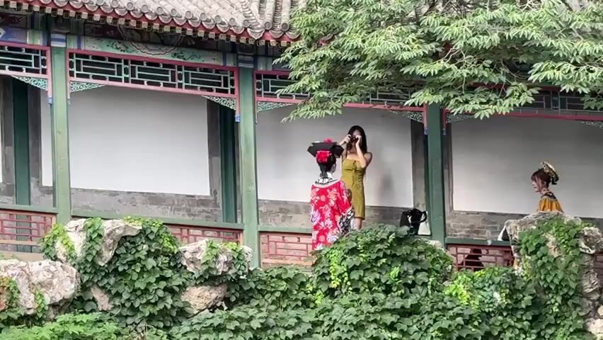 The video opens on a lush green landscape in front of a traditional Chinese building with ornate architecture. A person dressed in a red and gold traditional outfit stands on a ledge adorned with ivy. To their right, a person in a yellow dress is taking a photo or video with a phone. Further to the right, another person in a traditional outfit is visible, partially obscured by foliage. The camera then pans to the left, revealing a group of tourists gathered on a walkway in front of the building. The building itself features intricately painted beams, tiled roofs, and white walls. The overall scene depicts a tourist attraction with people dressed in both modern and traditional attire.