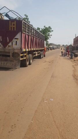 The video captures a daytime scene on a dusty, unpaved road in Kufena, Nigeria. Buildings, some appearing commercial or residential, line both sides of the road, interspersed with green trees.

A large truck, possibly a flatbed with an extended metal cage structure, is prominent on the left side of the road, either parked or moving slowly. Its side is painted with a distinctive yellow and brown pattern, featuring stylized yellow letters resembling "HA" or "HH," with a number "564" or "561" and "FG" visible below.

The primary activity shown is vehicular movement. Several individuals are seen riding motorcycles along the road. The camera initially focuses on the truck before panning to follow a man in a blue outfit riding a motorcycle, who is then followed by another motorcyclist. Other people are visible on the roadside, observing the passing traffic or engaged in their daily routines. The sky is clear, indicating sunny weather. The general atmosphere depicts typical street activity in a semi-urban or rural setting.