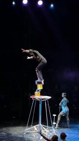 A circus performer balances precariously on a stack of cylindrical objects on a high platform. The performer, dressed in black, is bent over, with one leg extended. The platform is supported by a metal structure with chains. Another performer, with painted skin, stands on the stage below, holding a hoop. The stage is dark, with spotlights illuminating the performers. Several other figures are visible in the background, their faces obscured. The scene appears to be part of a live performance.