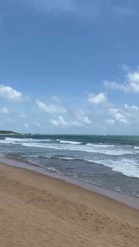 The video shows a sandy beach with gentle waves rolling onto the shore under a bright blue sky with scattered white clouds. The ocean is a mixture of turquoise and deep blue, and the sand is a light brown. There are no people or objects visible in the frame. The camera is positioned at a low angle, looking out towards the ocean. The scene is peaceful and serene.