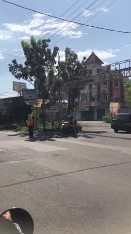 The video shows a street scene with a person in an orange vest standing near a road. A motorcycle with two people on it is passing by. There are trees, buildings, and overhead wires in the background. The sky is blue with some clouds. A person in an orange vest is standing near a white vehicle.