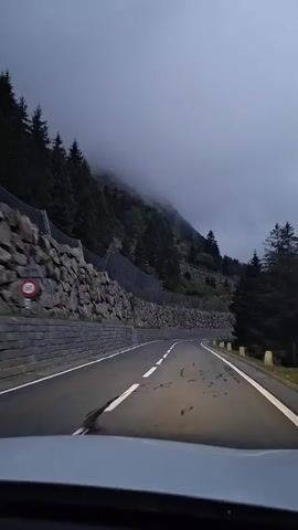 The video shows a point-of-view shot from inside a vehicle driving on a winding mountain road. The road has a grey asphalt surface with white lane markings. On the left side of the road, there is a steep embankment covered with a stone retaining wall topped with a metal fence. Evergreen trees are visible on the slope above the fence. The sky is overcast and foggy, with the mist obscuring the tops of the mountains. The right side of the road has a narrow paved shoulder and a small white marker post. The overall lighting suggests it is either early morning, late evening, or a very cloudy day.