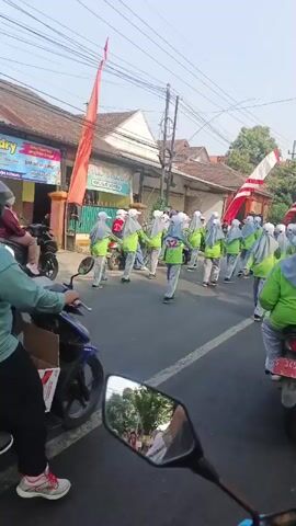 A group of people, appearing to be students wearing light green shirts, gray pants, and white hats, are walking down a street in what looks like a parade or procession. They are accompanied by motorcycles, one of which is being ridden by the videographer, providing a first-person perspective. Red and white flags are visible along the street, suggesting a celebratory event. The scene appears to be in a residential or commercial area with buildings and a traffic light visible.