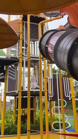 This is a video of a playground in Prigen, Indonesia. The playground features a large, multi-level structure made of yellow metal poles and white mesh. There are also some large, cylindrical slides that are part of the structure. The playground is decorated with cartoon images of farm animals, including a prominent image of a white cow with pink accents.

Several children are visible playing on the playground. One child in a red shirt and blue jeans is climbing the stairs. Another child, wearing a light-colored shirt and blue jeans, is also on the playground structure, as is a person in a light pink shirt and blue jeans. One child is partially visible inside one of the enclosed sections of the playground.

The scene appears to be outdoors, with trees and greenery visible in the background. The sky is blue with some clouds, suggesting it is daytime, likely a clear and pleasant day. The overall atmosphere seems playful and active, typical of a children's playground. There is no visible text in the video.