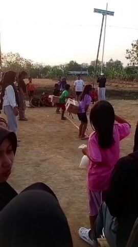 A group of children are playing a game outdoors. The game appears to involve carrying boxes. In the foreground, two girls are watching the game. One girl, wearing a pink shirt, holds a white bag. In the background, a group of children are participating in the game. Some children are carrying boxes, while others are watching. The ground is dirt and there are some trees and a volleyball net in the background. The sky is overcast.