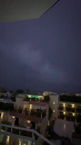 The video shows a dark, possibly night time, scene of buildings with lights on. There is a balcony in the foreground with a white railing. In the background, there are several buildings, some with illuminated windows and balconies. A swimming pool with green lights is visible in the center of the frame. The sky is dark and it appears to be raining lightly.