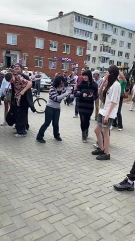 The video shows an outdoor scene with a group of young people gathered on a paved area. The background features a brick building with "Ozon" signage and apartment buildings. A few adults are also present, observing the scene. In the foreground, three young females are dancing or moving in a lively manner, holding onto each other. One of them has dark, short hair and is wearing a striped long-sleeved shirt and dark pants. Another has longer dark hair and is wearing a striped shirt with wide bands and dark leggings. The third, to the right, has brown hair and is wearing a red and black shirt and ripped denim jeans. To the far right, another young female in a white shirt and shorts is watching them. Behind the dancing group, a young girl is riding a bicycle, and several other people are visible, some standing and some walking. The overall atmosphere appears to be casual and energetic.