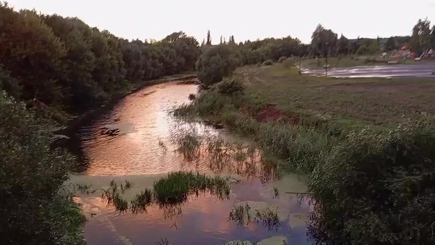 A slow aerial pan reveals a natural landscape at dusk. A narrow river flows through the center of the frame, its surface reflecting the warm, orange hues of the sunset-colored clouds above. Green vegetation lines the banks of the river, with dense trees forming a dark silhouette on the left side of the screen. On the right bank, a grassy area transitions into a paved surface, possibly a road or parking lot, with a few indistinct structures and equipment visible in the distance. The sky is a soft gradient of blue and pink, with scattered clouds catching the last light of the day. No people are visible in the video. The overall atmosphere is peaceful and serene.